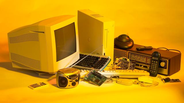 Elektroschrott wie ein alter PC und alte Handys vor gelben Hintergrund.