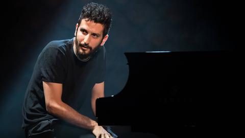 Vor schwarzen Hintergrund sitzt ein bärtiger junger Pianist am Flügel und richtet seinen Blick zur Seite.