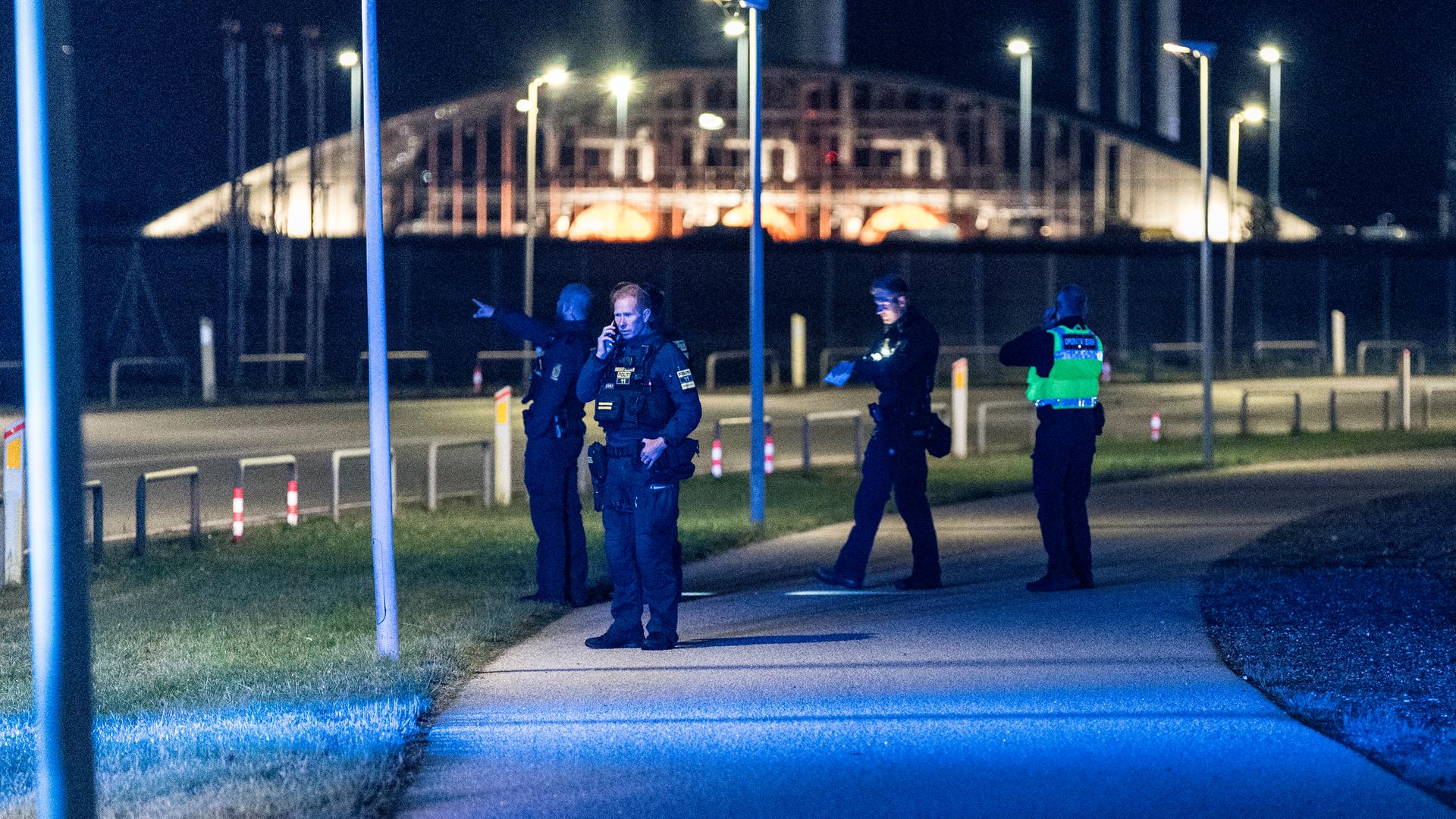 Polizisten stehen auf einer Straße am Flughafen von Kopenhagen. Einer spricht in ein Funkgerät, ein anderer zeigt in die Luft. Im Hintergrund sieht man ein größeres Gebäude.