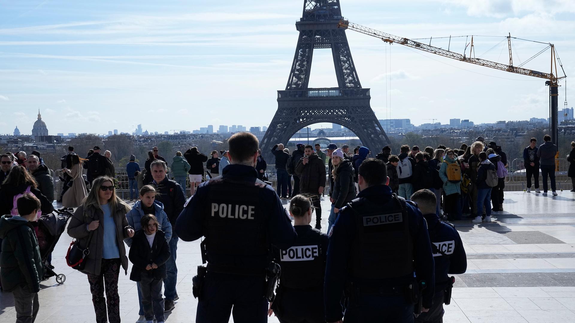Polizisten stehen auf dem Trocadero-Platz in Paris. Der Platz ist gut besucht. Im Hintergrund sieht man den Fuß des Eiffelturms.