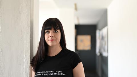  Die Schriftstellerin Mareike Fallwickl steht in einem Flur mit weißen Wänden. Sie trägt ein schwarzes T-Shirt mit weißer Aufschrift.