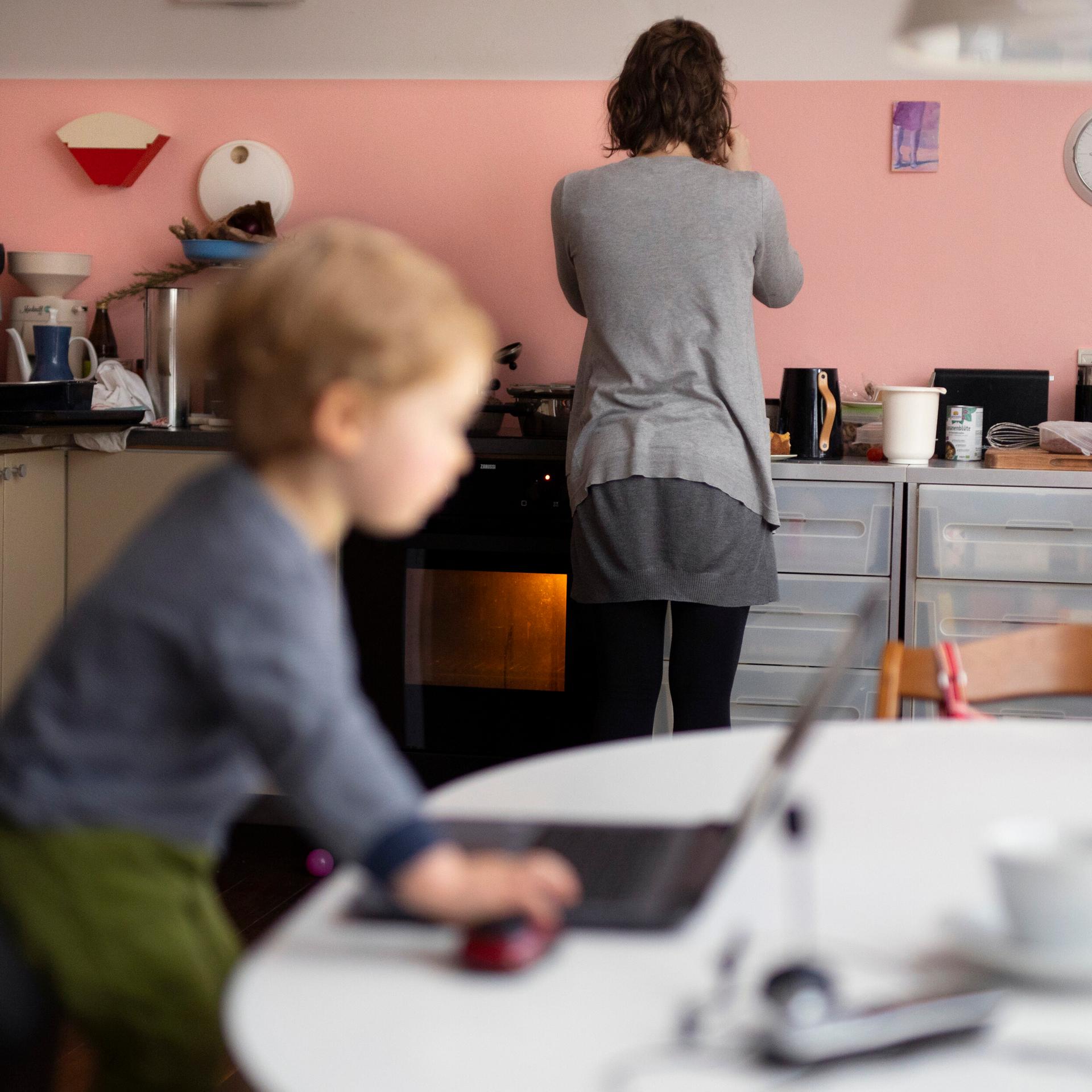 Das Foto zeigt eine Frau in der Küche, ihr Kleinkind steht auf einem Stuhl vor einem Computer