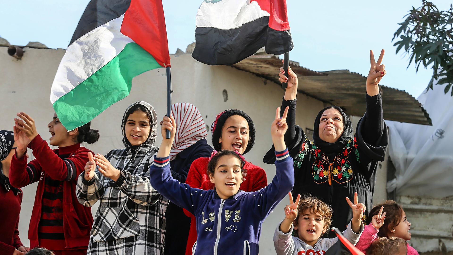 Kinder und Frauen mit Kopftuch stehen zusammen und feiern. Einige applaudieren, andere machen das "Peace"-Zeiche. Sie schwänken palästinensiche Flaggen.