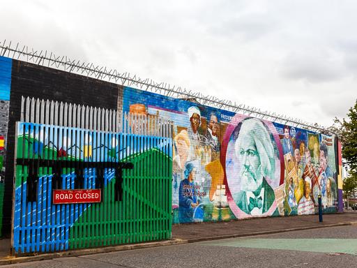Wandbemalungen in der Northumberland Street, nahe der Falls Road in Belfast. Davor ein Tor mit der Aufschrift "Road closed", das eine sogenannte Peace Line (Friedenslinie) zur Trennung von Konfliktparteien darstellt. Abends wird das Tor geschlossen. 