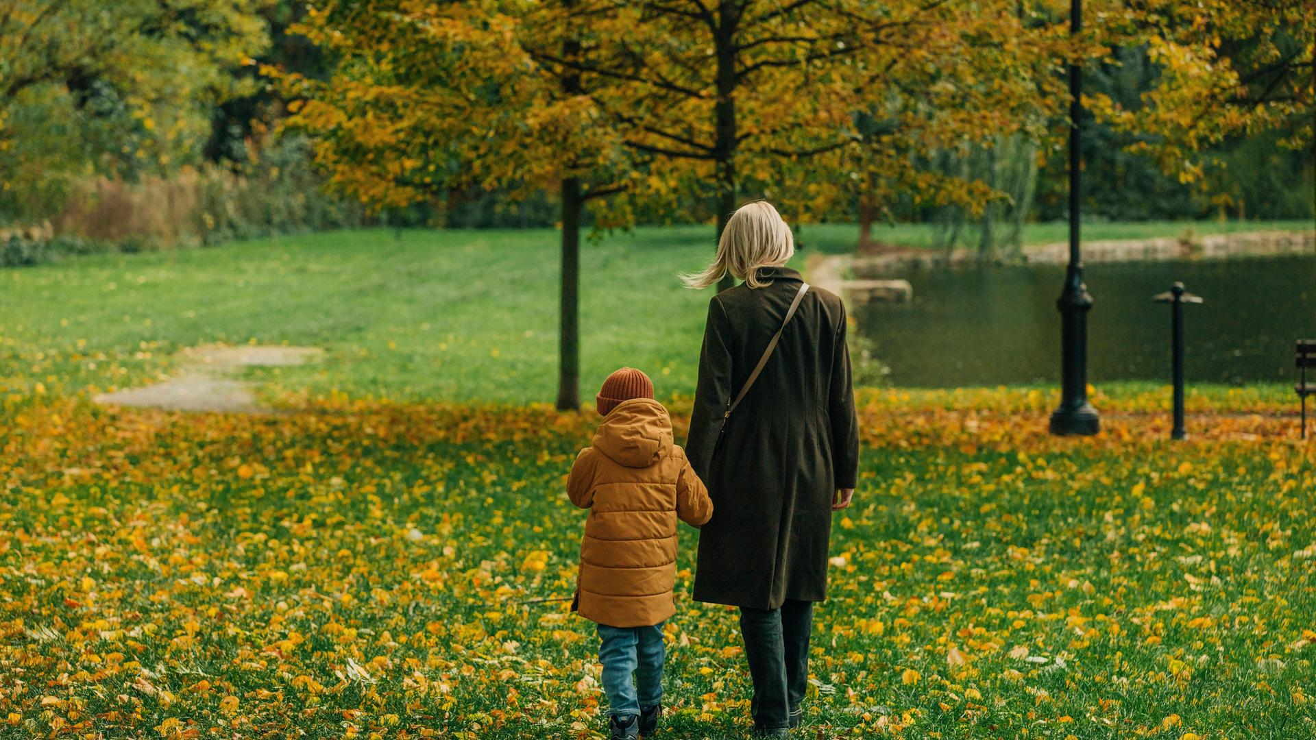 Eine Frau mit blonden Haaren in Herbstmantel läuft durch eine grüne Wiese in einem Park. An der Hand führt sie ihr Kind in gelber Jacke. 