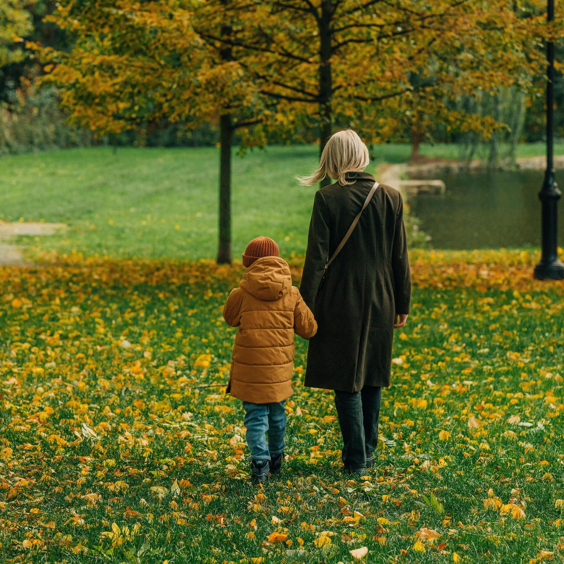 Eine Frau mit blonden Haaren in Herbstmantel läuft durch eine grüne Wiese in einem Park. An der Hand führt sie ihr Kind in gelber Jacke. 