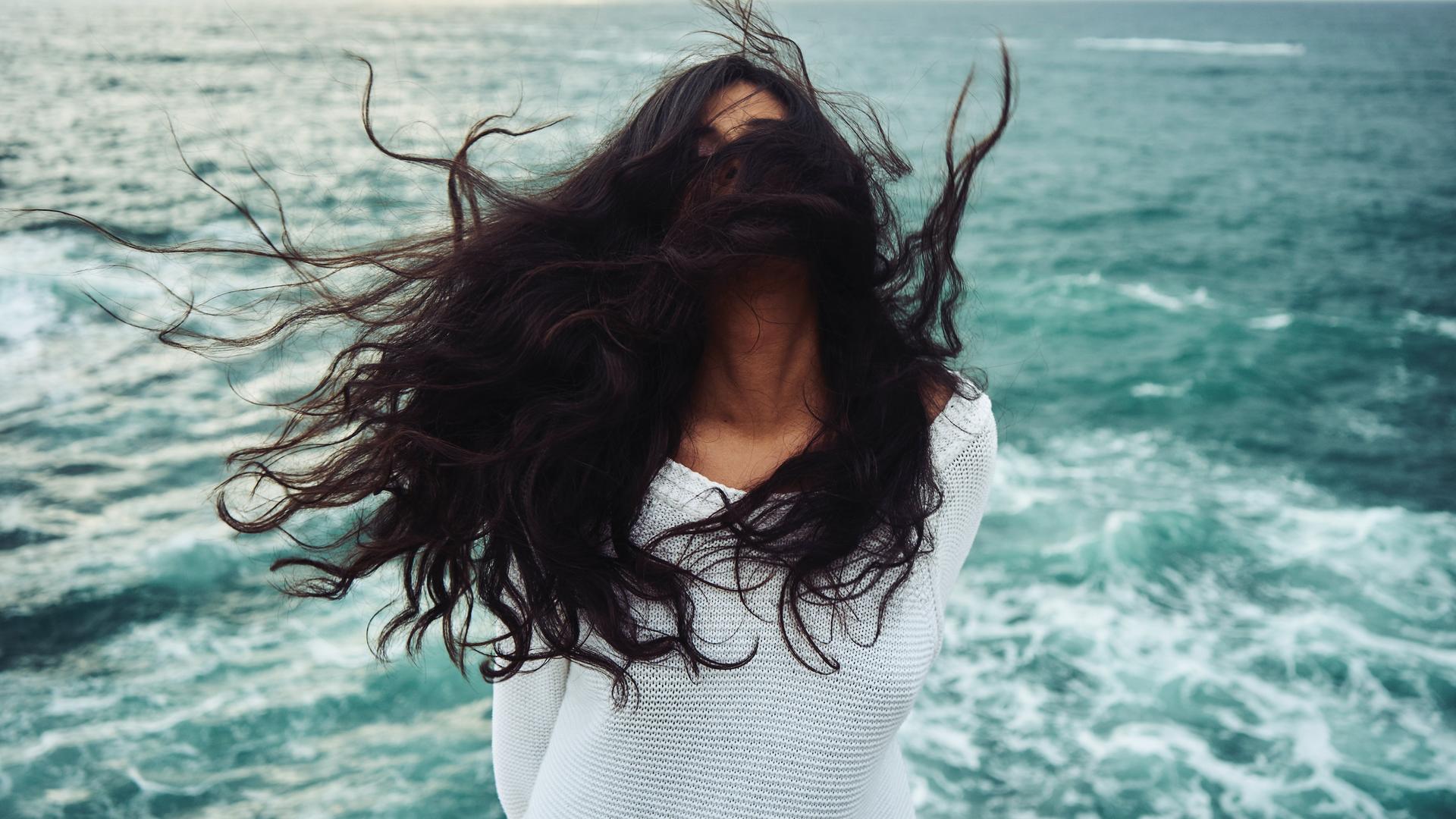 Eine Frau mit dunklen langen Jahren steht im Wind am Meer, ihre Haare sind so zerzaust, dass man das Gesicht nicht erkennen kann.