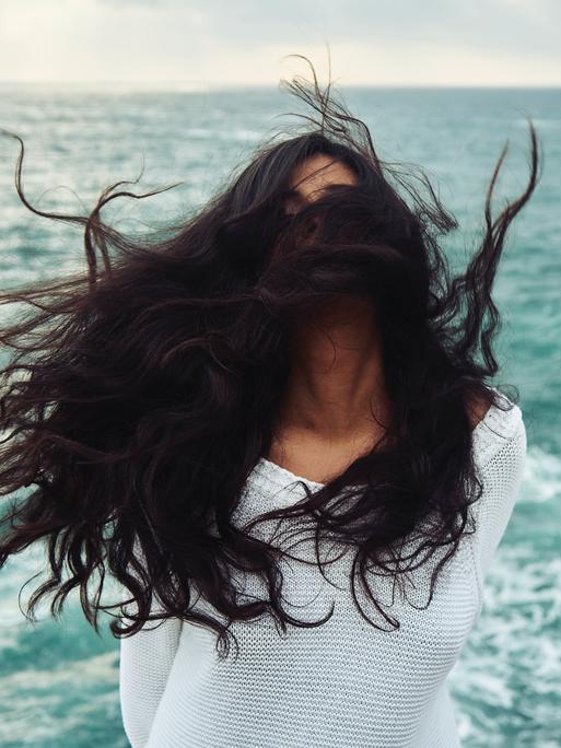 Eine Frau mit dunklen langen Jahren steht im Wind am Meer, ihre Haare sind so zerzaust, dass man das Gesicht nicht erkennen kann.