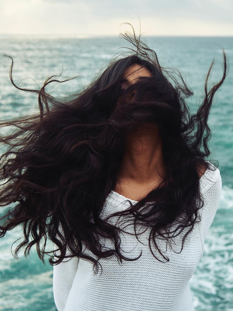 Eine Frau mit dunklen langen Jahren steht im Wind am Meer, ihre Haare sind so zerzaust, dass man das Gesicht nicht erkennen kann. Eine Frau mit dunklen langen Jahren steht im Wind am Meer, ihre Haare sind so zerzaust, dass man das Gesicht nicht erkennen kann.