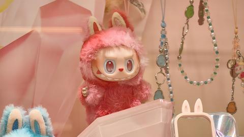 Eine kleine, grinsende, rosa Labubu-Puppe, umgeben von viel Bling-Bling. Sie zieht eine verschmitzt-bösartige Fratze. 
