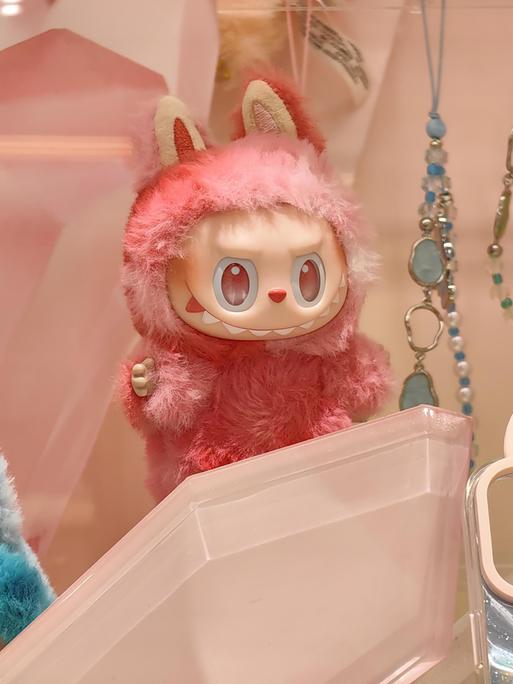 Eine kleine, grinsende, rosa Labubu-Puppe, umgeben von viel Bling-Bling. Sie zieht eine verschmitzt-bösartige Fratze. 