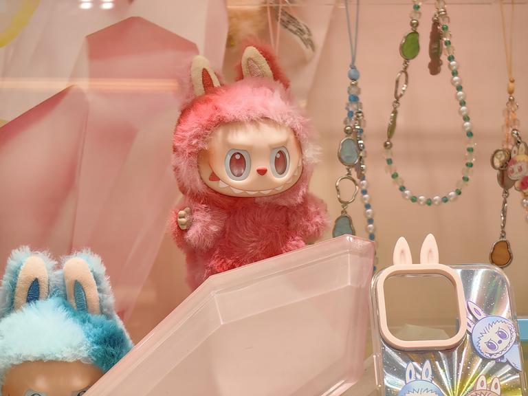 Eine kleine, grinsende, rosa Labubu-Puppe, umgeben von viel Bling-Bling. Sie zieht eine verschmitzt-bösartige Fratze. 