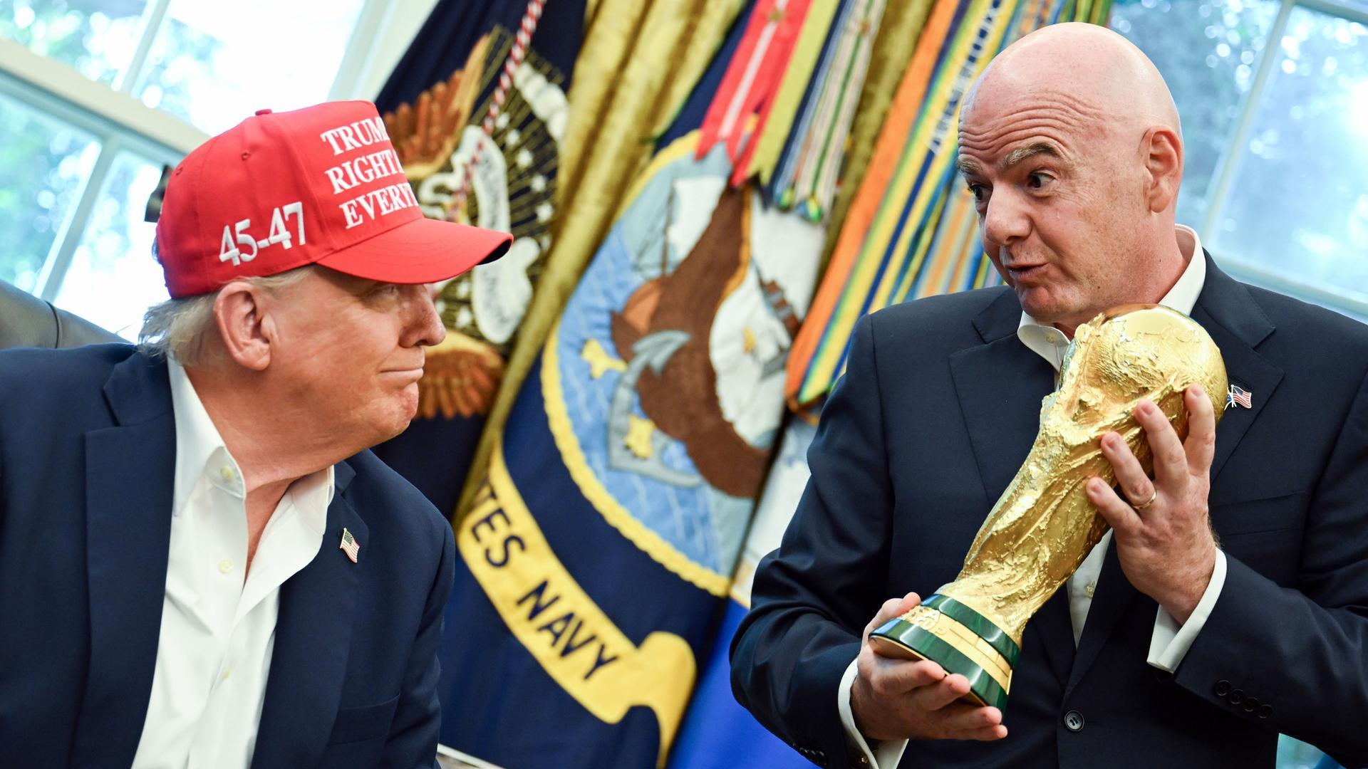 FIFA-Präsident Gianni Infantino hält im Oval Offive den goldenen Fußball-WM-Pokal in den Händen und schaut dabei auf den sitzenden US-Präsident Donald Trump herab. Der trägt eine rote Kappe und grinst zurück. 