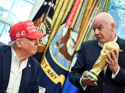 FIFA-Präsident Gianni Infantino hält im Oval Offive den goldenen Fußball-WM-Pokal in den Händen und schaut dabei auf den sitzenden US-Präsident Donald Trump herab. Der trägt eine rote Kappe und grinst zurück. FIFA-Präsident Gianni Infantino hält im Oval Offive den goldenen Fußball-WM-Pokal in den Händen und schaut dabei auf den sitzenden US-Präsident Donald Trump herab. Der trägt eine rote Kappe und grinst zurück.