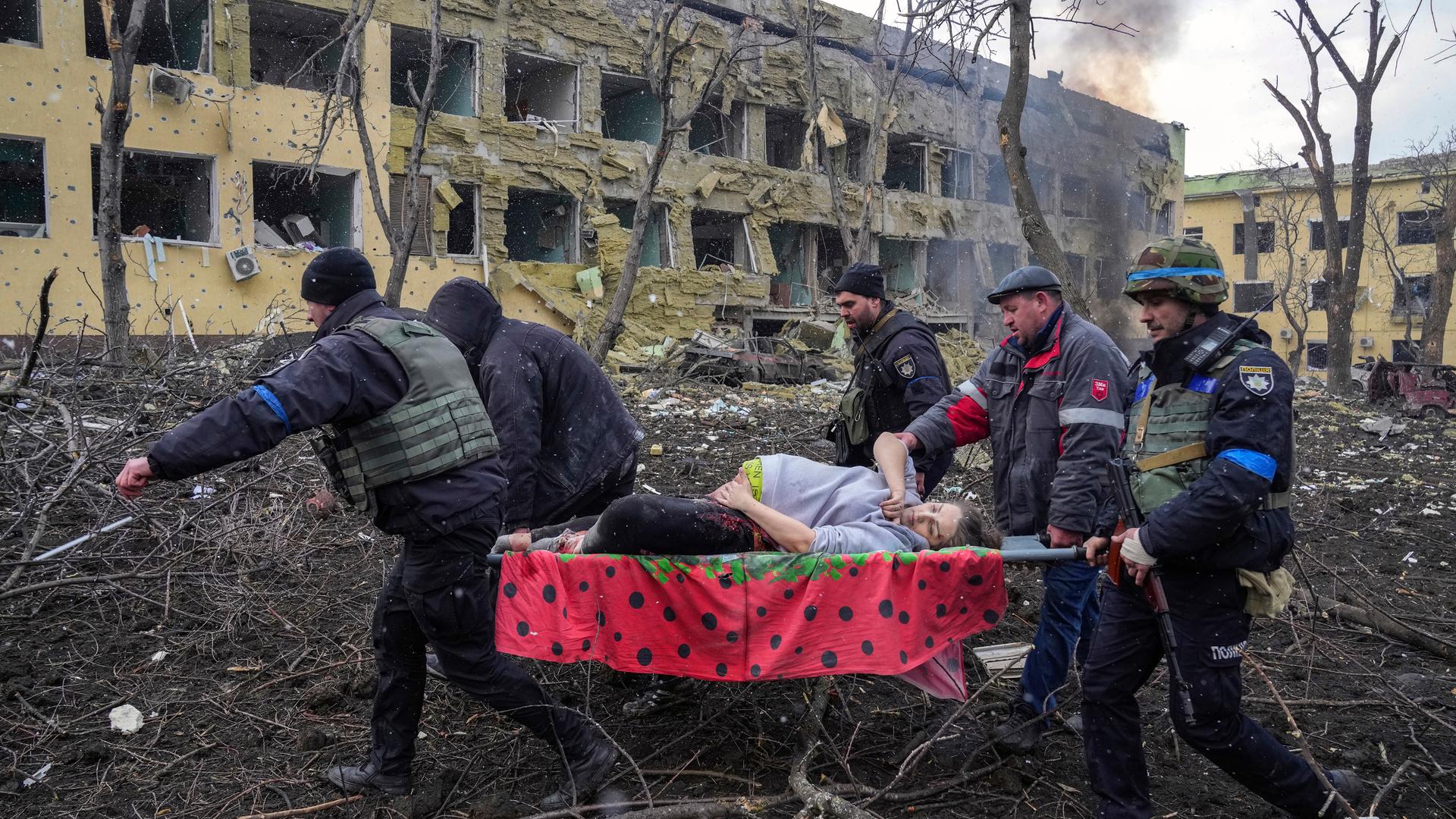 Fünf Männer, darunter ein Soldat, transportieren eine schwangere Frau auf einer Trage an dem zerstörten Gebäude vorbei durch einen ebenso zerstören Garten. Sie hält ihre Hand auf eine blutende Wunde am Bauch.