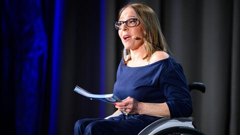 Miriam Labus sitzt in ihrem Rollstuhl auf einer Bühne. Sie trägt ein Mikrofon am Kopf und spricht. Miriam Labus sitzt in ihrem Rollstuhl auf einer Bühne. Sie trägt ein Mikrofon am Kopf und spricht.