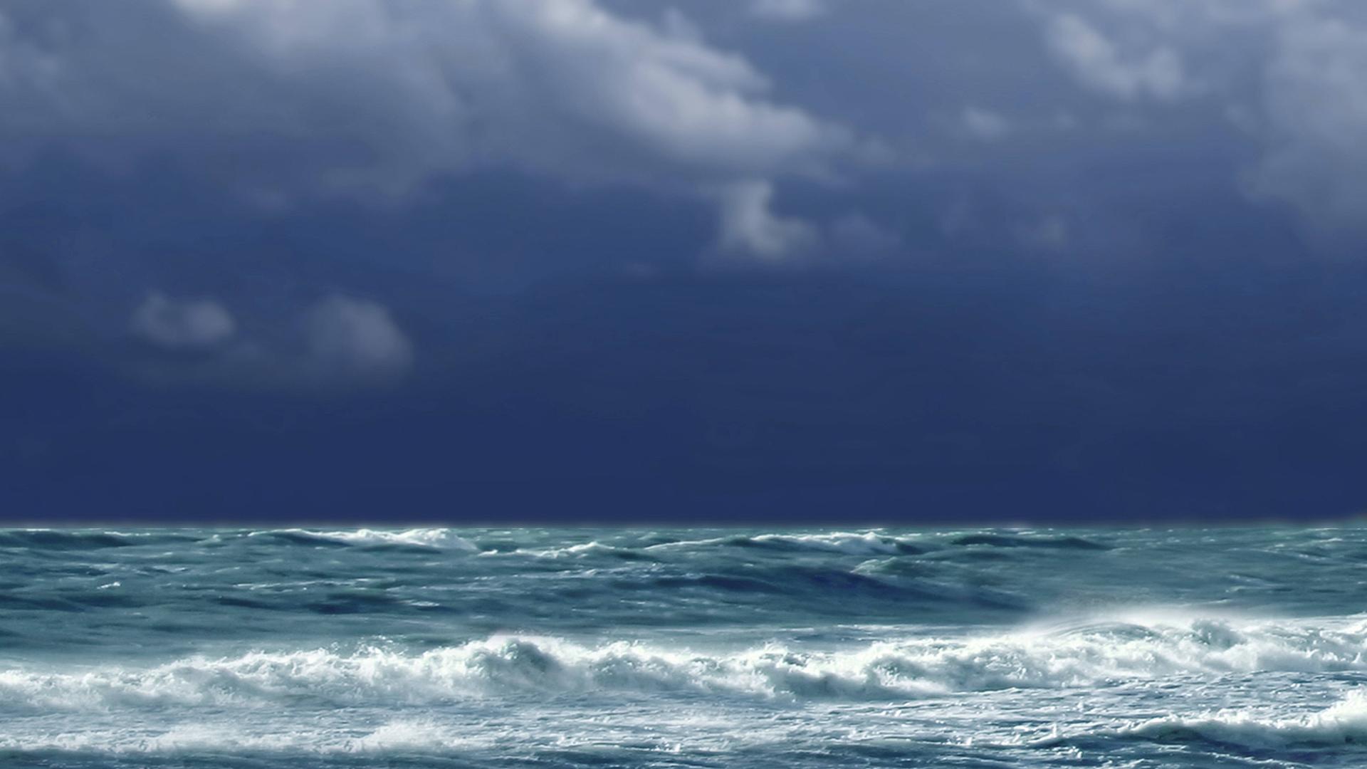 Über der aufgewühlten See sieht man einen  sehr dunklen Himmel. Am vorderen Bildrand ein Kiesstrand.