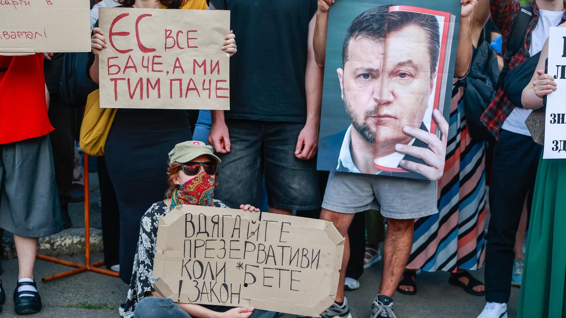 Protestierende halten Plakate hoch, eines davon zeigt eine Montage des Gesichts von Präsident Wolodymyr Selenskyj mit dem des früheren ukrainischen Präsidenten Viktor Janukowitsch