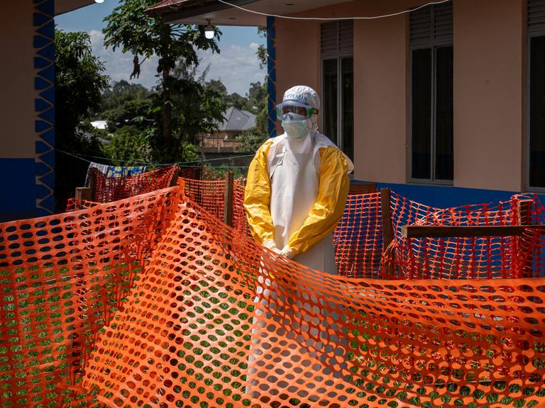 Eine Person in Schutzkleidung steht vor einer durch Zäune abgesperrten Klinik. Eine Person in Schutzkleidung steht vor einer durch Zäune abgesperrten Klinik.