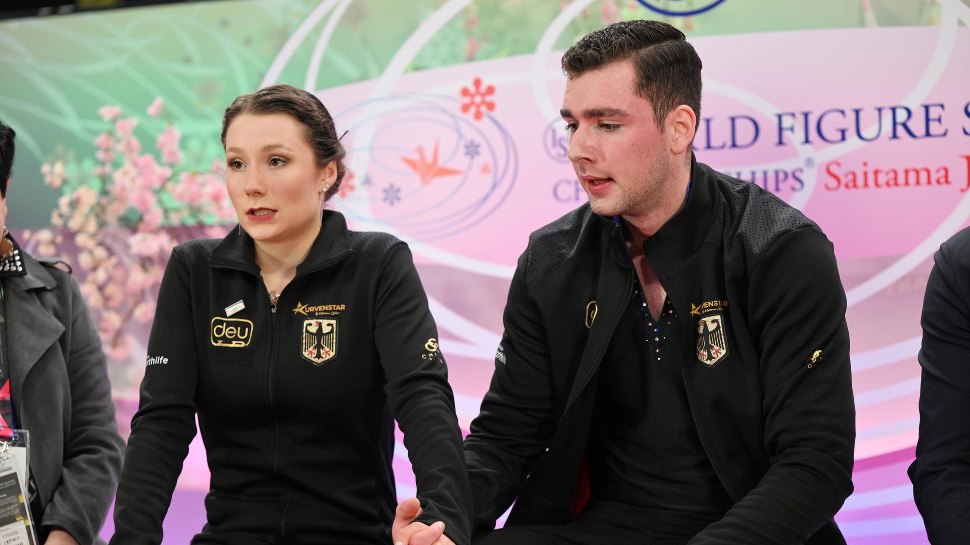Das Eiskunstlaufpaar Annika Hocke und Robert Kunkel warten bei den Weltmeisterschaften 2023 in Saitama auf ihre Wertnoten. Dabei halten sie sich an den Händen. 