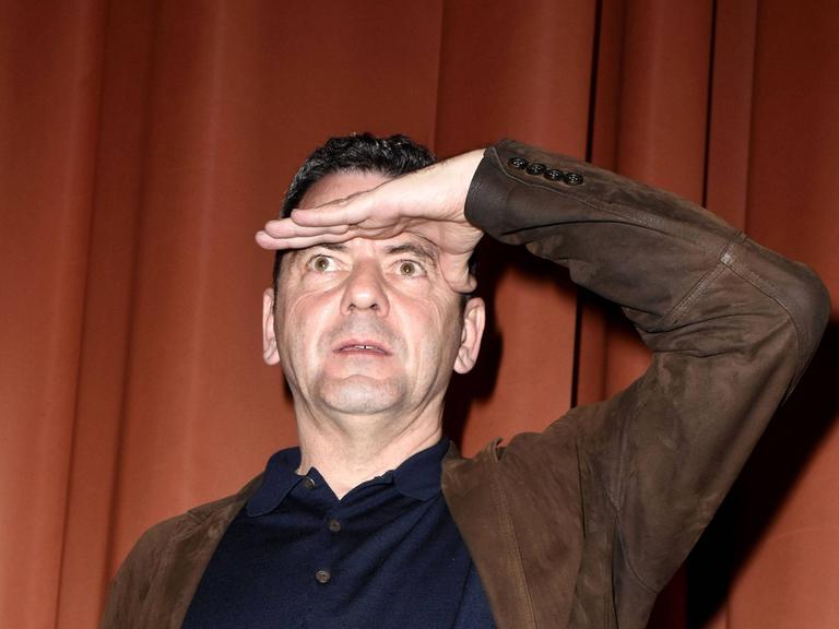 Christian Petzold steht vor einem Vorhang auf einer Bühne und schaut mit einer Handfläche an die Stirn gelegt in die Ferne.  