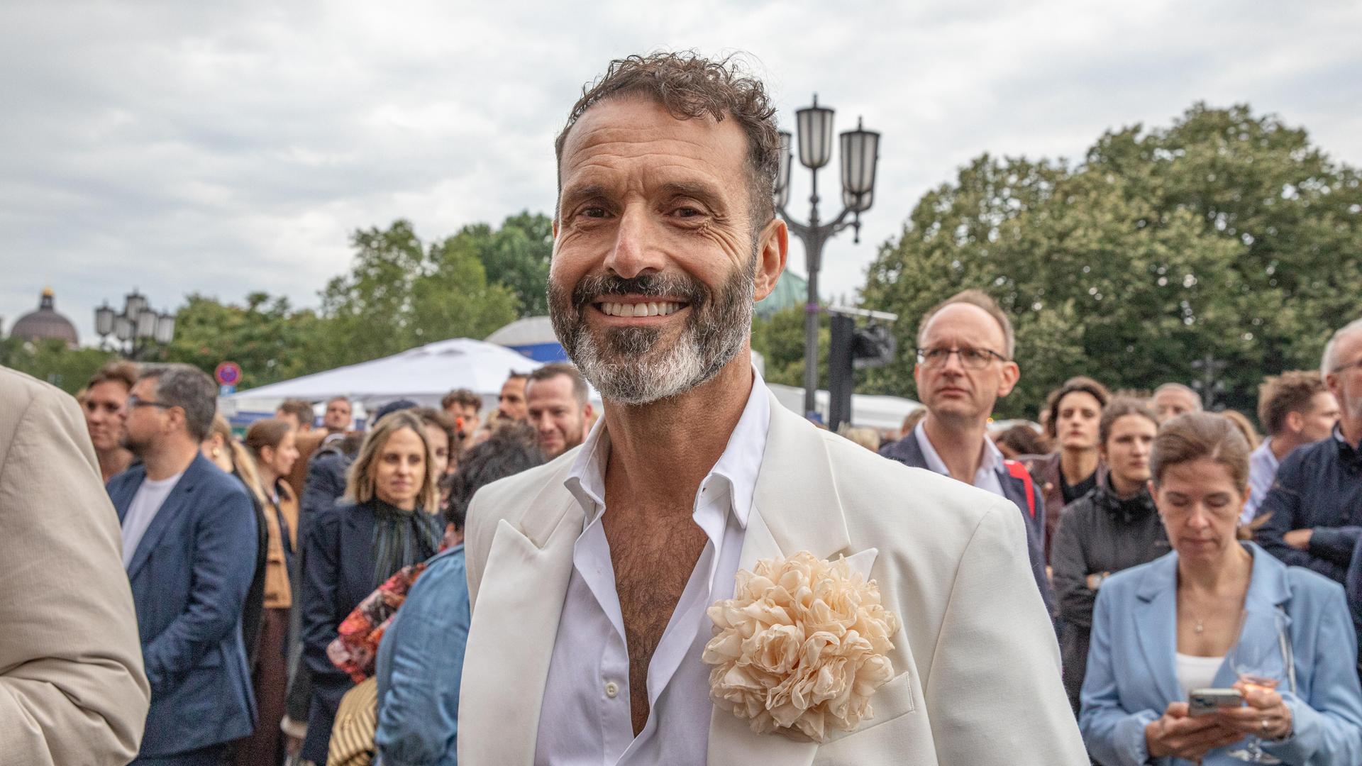 Ein Mann mit hellem Anzug und weißem Hemd lächelt in die Kamera bei einem Fest. Es ist der Queer-Beauftragte Berlin, Alfonso Pantisano.