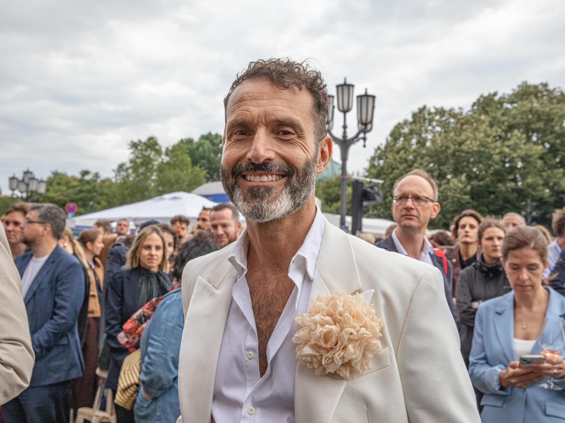 Ein Mann mit hellem Anzug und weißem Hemd lächelt in die Kamera bei einem Fest. Es ist der Queer-Beauftragte Berlin, Alfonso Pantisano.