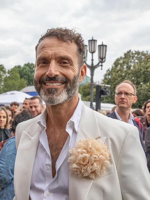 Ein Mann mit hellem Anzug und weißem Hemd lächelt in die Kamera bei einem Fest. Es ist der Queer-Beauftragte Berlin, Alfonso Pantisano.