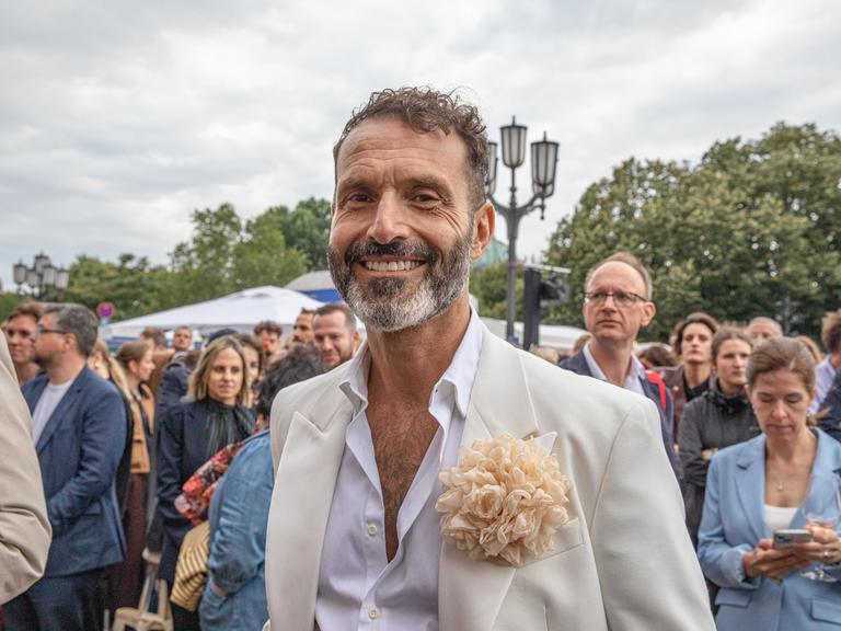 Ein Mann mit hellem Anzug und weißem Hemd lächelt in die Kamera bei einem Fest. Es ist der Queer-Beauftragte Berlin, Alfonso Pantisano.