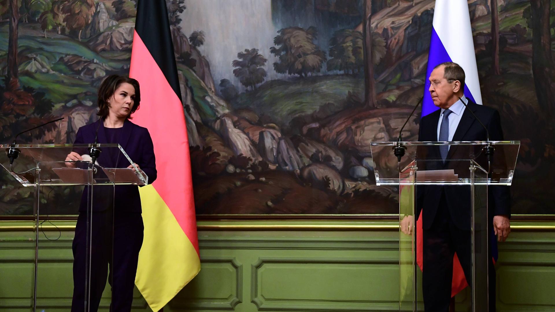Baerbock und Lawrow stehen an Pulten mit Mikrofonen vor einem großen Gemälde. Hinter den Pulten stehen die Fahnen ihrer Länder. 