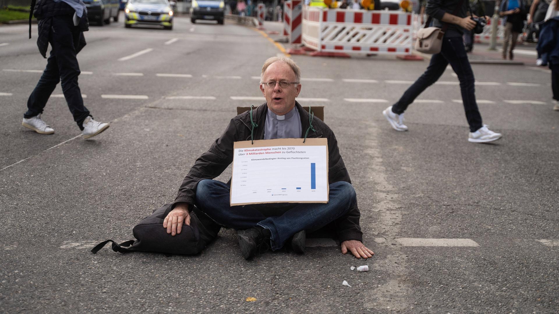 Blockade von "Scientists Rebellion" in München: Der Jesuiten-Pater Jörg Alt sitzt mit einem Schild mit der Aufschrift "Die Klimakatastrophe macht bis 2070 über 3 Milliarden Menschen zu Geflüchteten" um den Hals auf der Straße.
