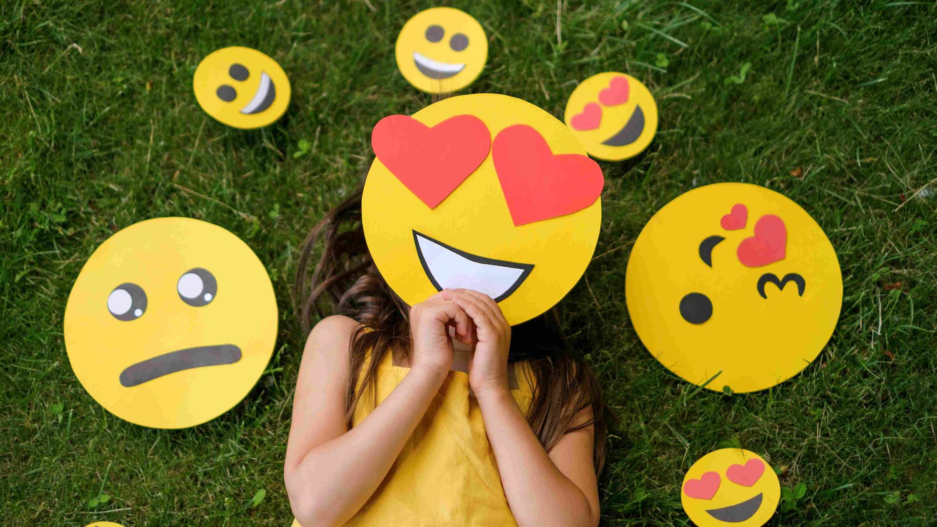 Ein Mädchen liegt auf einer Wiese, vor das Gesicht hält es eine Emoji-Maske mit Herzchen-Augen. Um den Kopf herum liegen diverse andere Emojis, die andere Gefühle ausdrücken. Ein Mädchen liegt auf einer Wiese, vor das Gesicht hält es eine Emoji-Maske mit Herzchen-Augen. Um den Kopf herum liegen diverse andere Emojis, die andere Gefühle ausdrücken.
