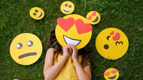 Ein Mädchen liegt auf einer Wiese, vor das Gesicht hält es eine Emoji-Maske mit Herzchen-Augen. Um den Kopf herum liegen diverse andere Emojis, die andere Gefühle ausdrücken.
