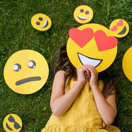 Ein Mädchen liegt auf einer Wiese, vor das Gesicht hält es eine Emoji-Maske mit Herzchen-Augen. Um den Kopf herum liegen diverse andere Emojis, die andere Gefühle ausdrücken.