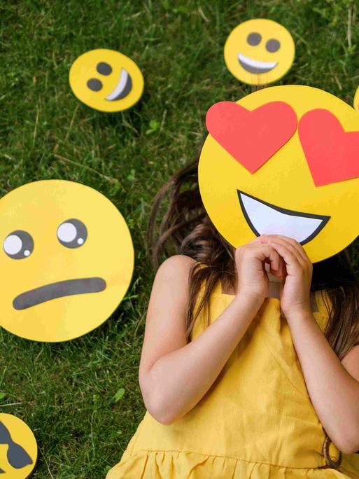 Ein Mädchen liegt auf einer Wiese, vor das Gesicht hält es eine Emoji-Maske mit Herzchen-Augen. Um den Kopf herum liegen diverse andere Emojis, die andere Gefühle ausdrücken.