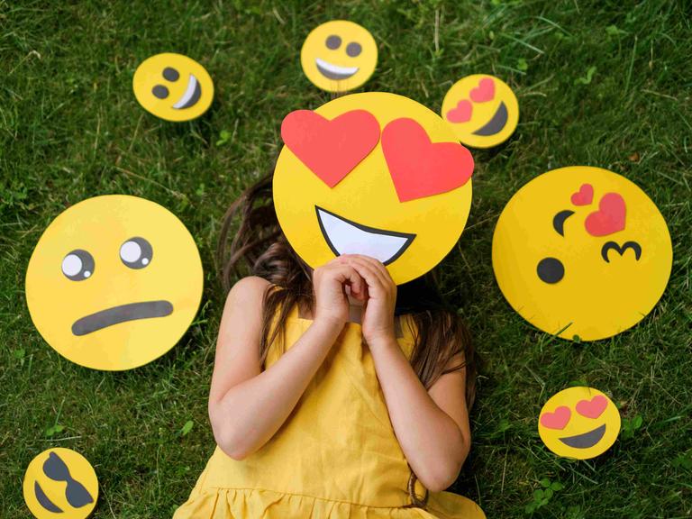 Ein Mädchen liegt auf einer Wiese, vor das Gesicht hält es eine Emoji-Maske mit Herzchen-Augen. Um den Kopf herum liegen diverse andere Emojis, die andere Gefühle ausdrücken. Ein Mädchen liegt auf einer Wiese, vor das Gesicht hält es eine Emoji-Maske mit Herzchen-Augen. Um den Kopf herum liegen diverse andere Emojis, die andere Gefühle ausdrücken.