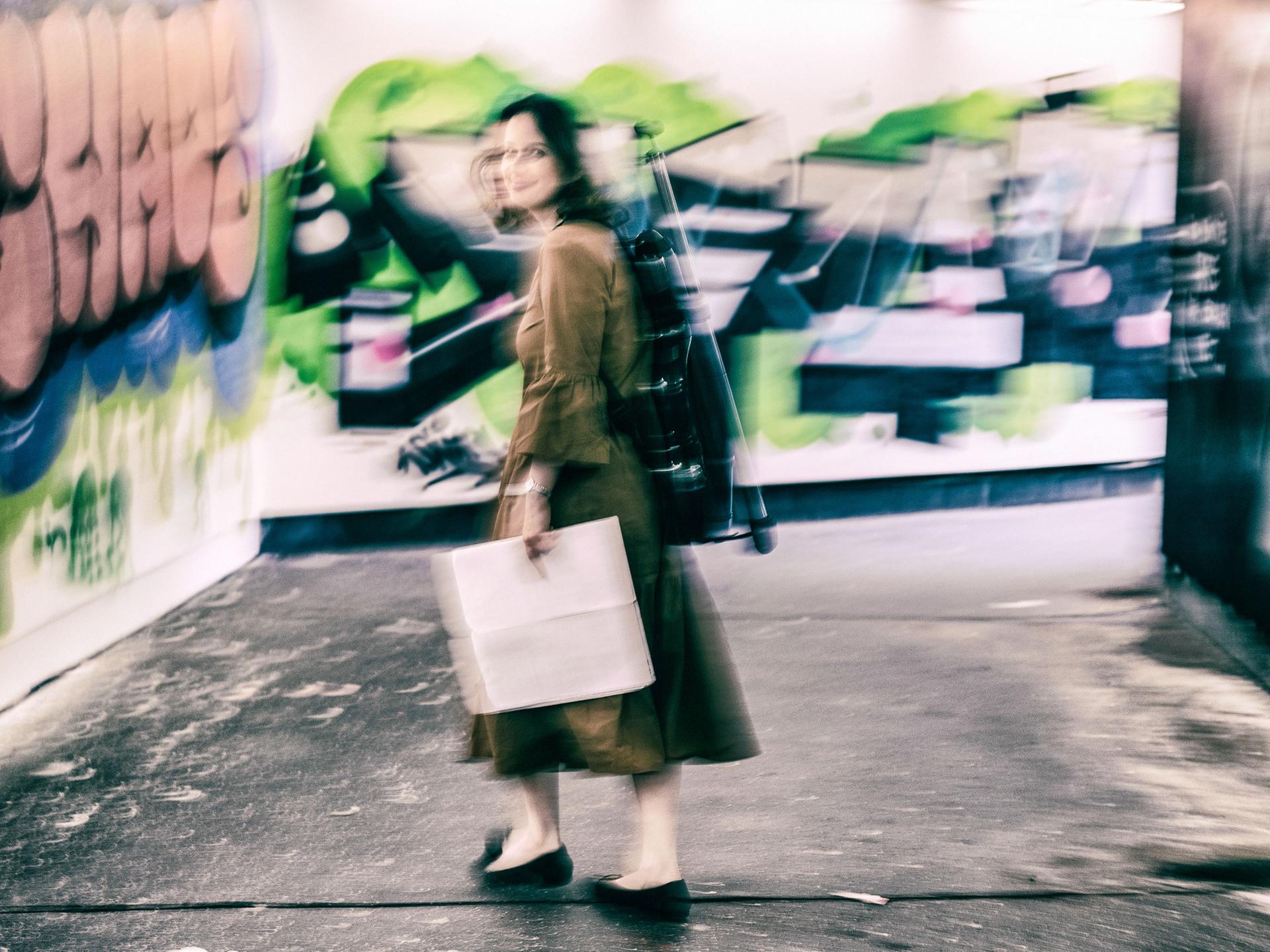 Patrica Kopatchinskaja steht in einer Unterführung mit Graffiti. Sie trägt ihren Geigenrucksack auf dem Rücken und hält Noten in der Hand. Das Bild fängt ihre Bewegung mit ein.