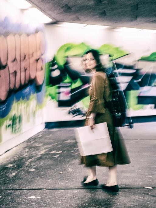 Patrica Kopatchinskaja steht in einer Unterführung mit Graffiti. Sie trägt ihren Geigenrucksack auf dem Rücken und hält Noten in der Hand. Das Bild fängt ihre Bewegung mit ein.