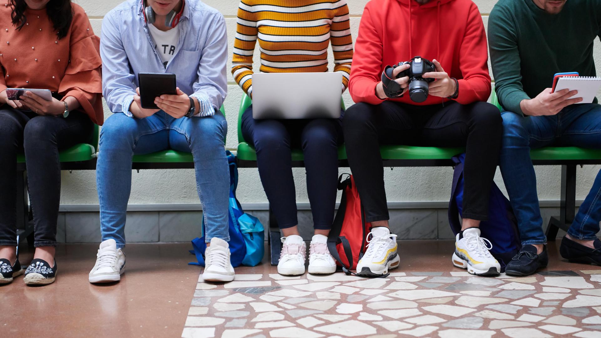 Mehrere junge Menschen sitzen auf einem Flur. Sie halten digitale Endgeräte, eine Fotokamera oder einen Block in der Hand.