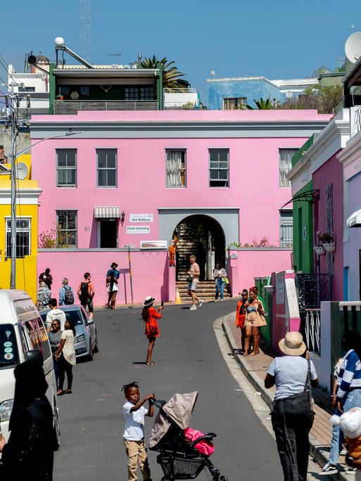 Bunte Häuser stehen im Stadtviertel Bo-Kaap in Kapstadt/Südafrika, viele Menschen laufen auf der Straße. Der Stadtteil entstand als Siedlung von Kapmalaien, die bis heute die Mehrheit der Bewohner stellen. 
