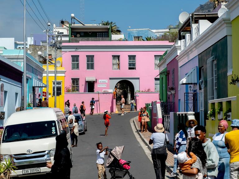 Bunte Häuser stehen im Stadtviertel Bo-Kaap in Kapstadt/Südafrika, viele Menschen laufen auf der Straße. Der Stadtteil entstand als Siedlung von Kapmalaien, die bis heute die Mehrheit der Bewohner stellen. 