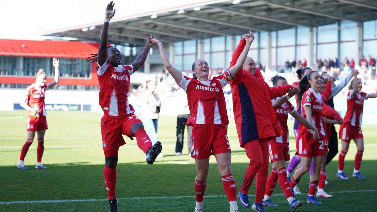 Mehrere Fußballerinnen mit rot-weißen Trikots und Sporthosen jubeln gemeinsam mit hochgereckten Armen vor der Fankurve.
