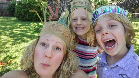 Blonde Mutter und zwei blonde Kinder machen Faxen mit Papierkronen im Garten.