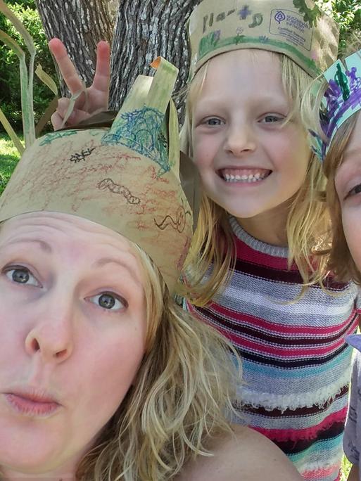 Blonde Mutter und zwei blonde Kinder machen Faxen mit Papierkronen im Garten.