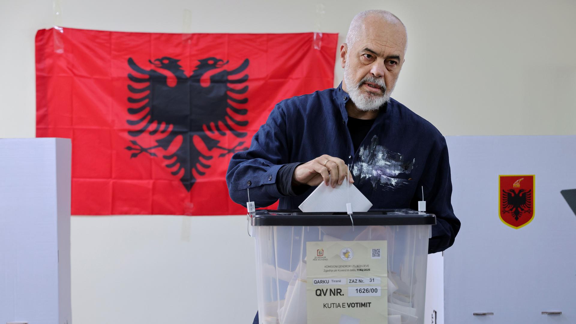 Rama steckt seinen Stimmzettel mit ernster Mine in eine Wahlurne aus Plastik. Dahinter eine Wand mit der rot-schwarzen Flagge Albaniens. Rama steckt seinen Stimmzettel mit ernster Mine in eine Wahlurne aus Plastik. Dahinter eine Wand mit der rot-schwarzen Flagge Albaniens.