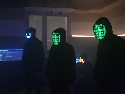 Schwarze Silhouetten von vermeintlich jungen Männern mit Kapuzenpullis in einem abgedunkelten Raum. Ihre Gesichter sind mit digitalen Codes unkenntlich gemacht. 