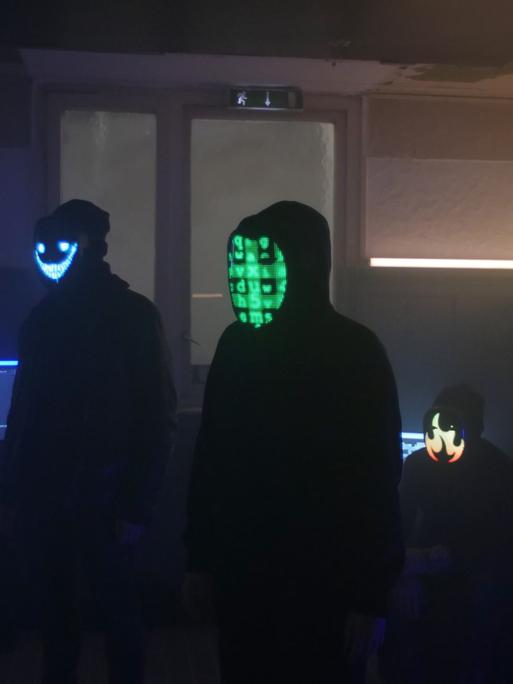 Schwarze Silhouetten von vermeintlich jungen Männern mit Kapuzenpullis in einem abgedunkelten Raum. Ihre Gesichter sind mit digitalen Codes unkenntlich gemacht. 
