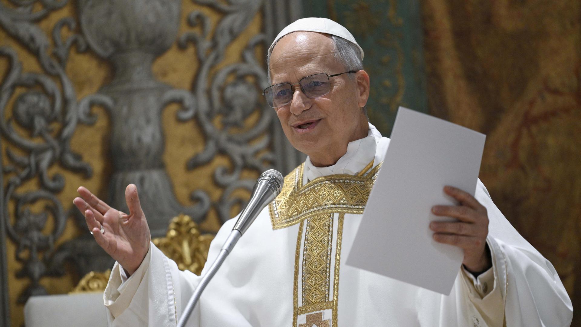 Papst Leo spricht und hebt die Arme. In einer Hand hält er Papiere. Er trägt eine weiße Kopfbedeckung und ein weiß-goldenes Gewand. Papst Leo spricht und hebt die Arme. In einer Hand hält er Papiere. Er trägt eine weiße Kopfbedeckung und ein weiß-goldenes Gewand.
