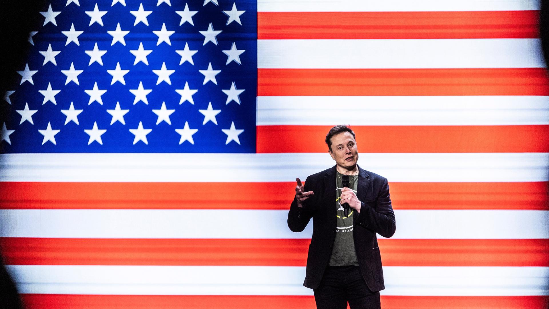 Der Unternehmer Elon Musk steht auf einer Bühne. Im Hintergrund eine große Flagge der Vereinigten Staaten von Amerika.