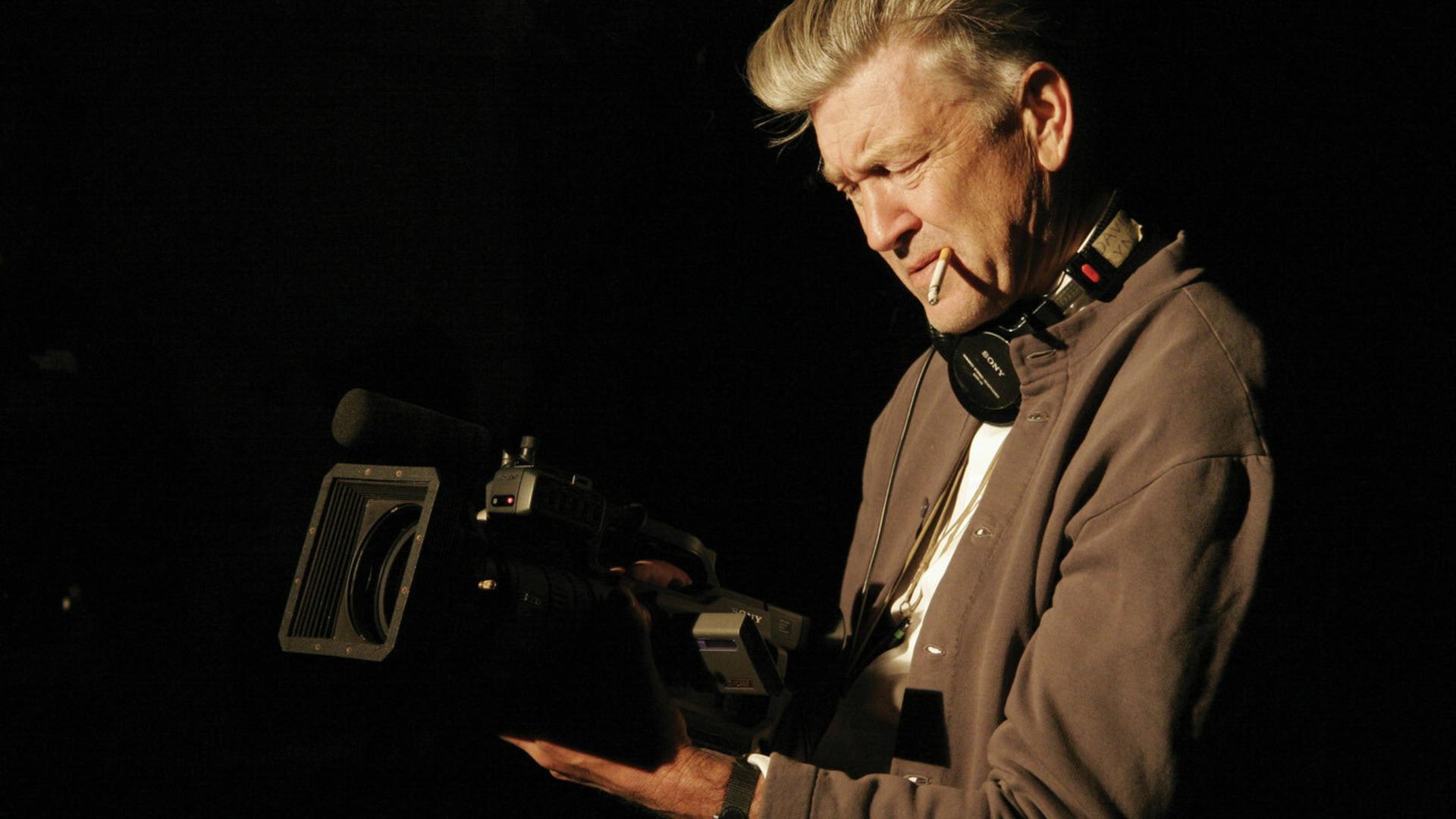 David Lynch hält konzentriert eine laufende Kamera in beiden Händen, eine Zigarette im Mundwinkel und Kopfhörer um den Hals